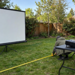 How Far Should Your Projector Be From the Screen Outdoors in 2026 Projector on table behind lawn chairs projecting onto outdoor screen at night