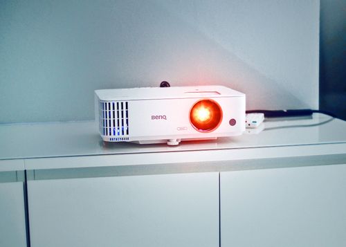 White projector on a cabinet with glowing lens - How To Choose A Projector For Home Theater 2026